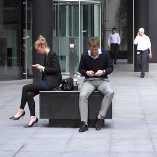 Two employees using their mobiles sat on a plinth outside