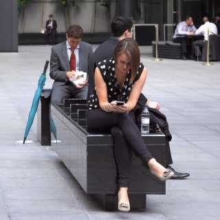 Two employees in business attire using their phones on a bench outside an office
